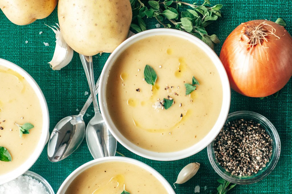 Kartoffelsuppe in Schüssel, daneben Löffel, Zwiebeln, Kartoffeln und Pfeffer